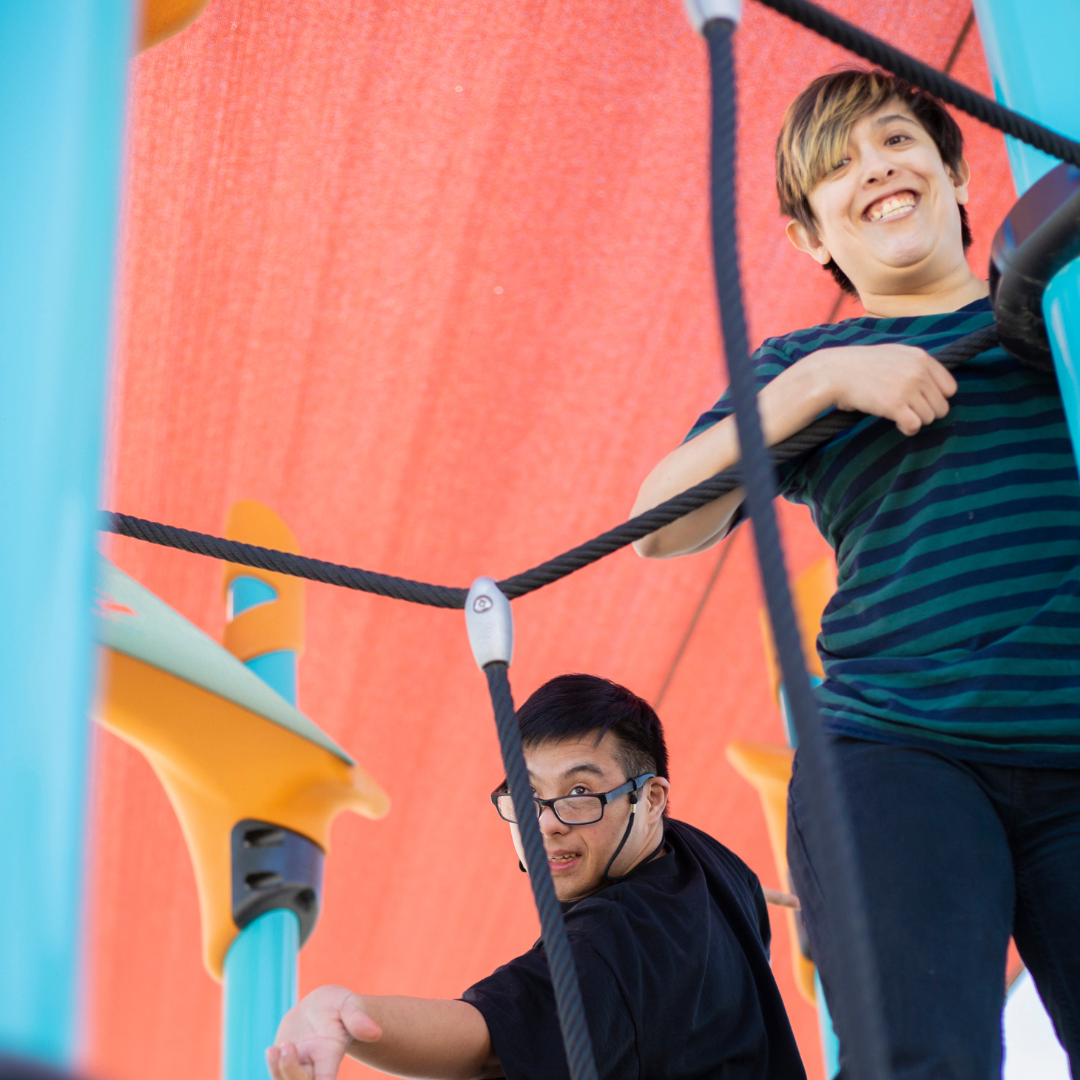Two people smiling on a playground with bright blue equipment and an orange overhead canopy.