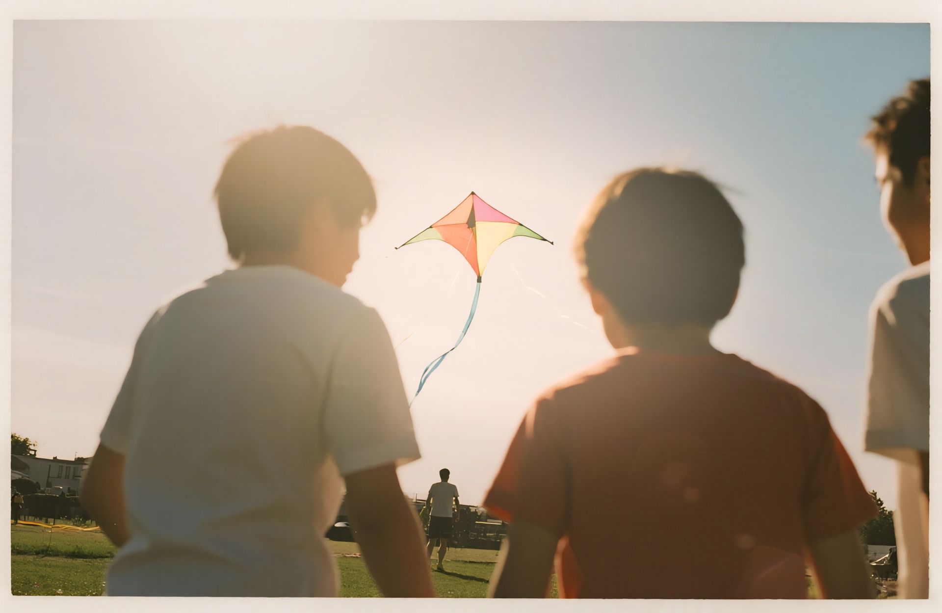 Three people stand outdoors at sunset, watching a colorful diamond-shaped kite flying in the sky.