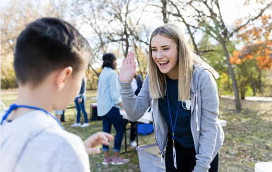 A volunteer high-fives a child outdoors at a park, both smiling and interacting during a community event.