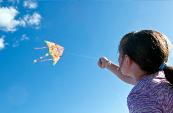 A person with hair tied back in a purple shirt reaches up to fly a colorful kite against a bright blue sky with clouds.