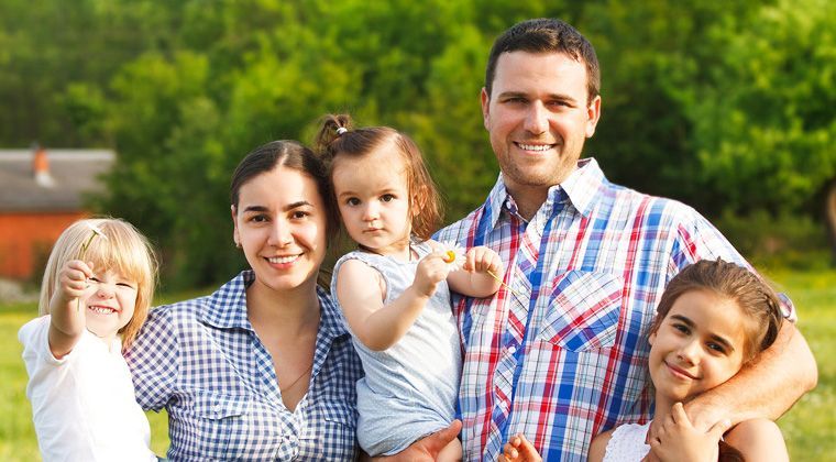 A family of five smiles together in a sunny, green outdoor setting.