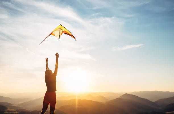 A person stands on a mountain peak at sunset, arms raised toward a yellow kite flying in the sky.