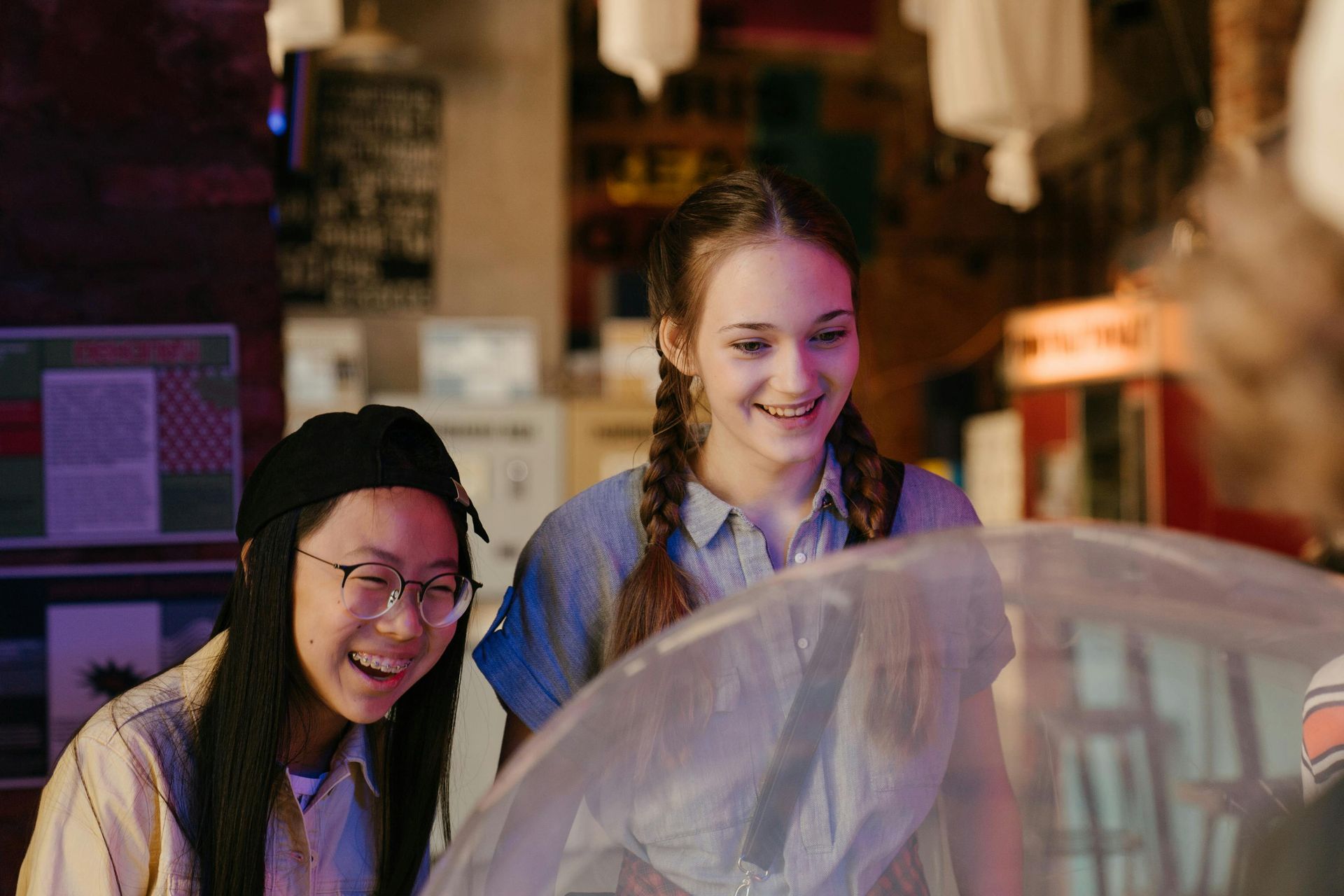 Two smiling people look toward a counter in a dimly lit indoor space, seen through a translucent foreground element.