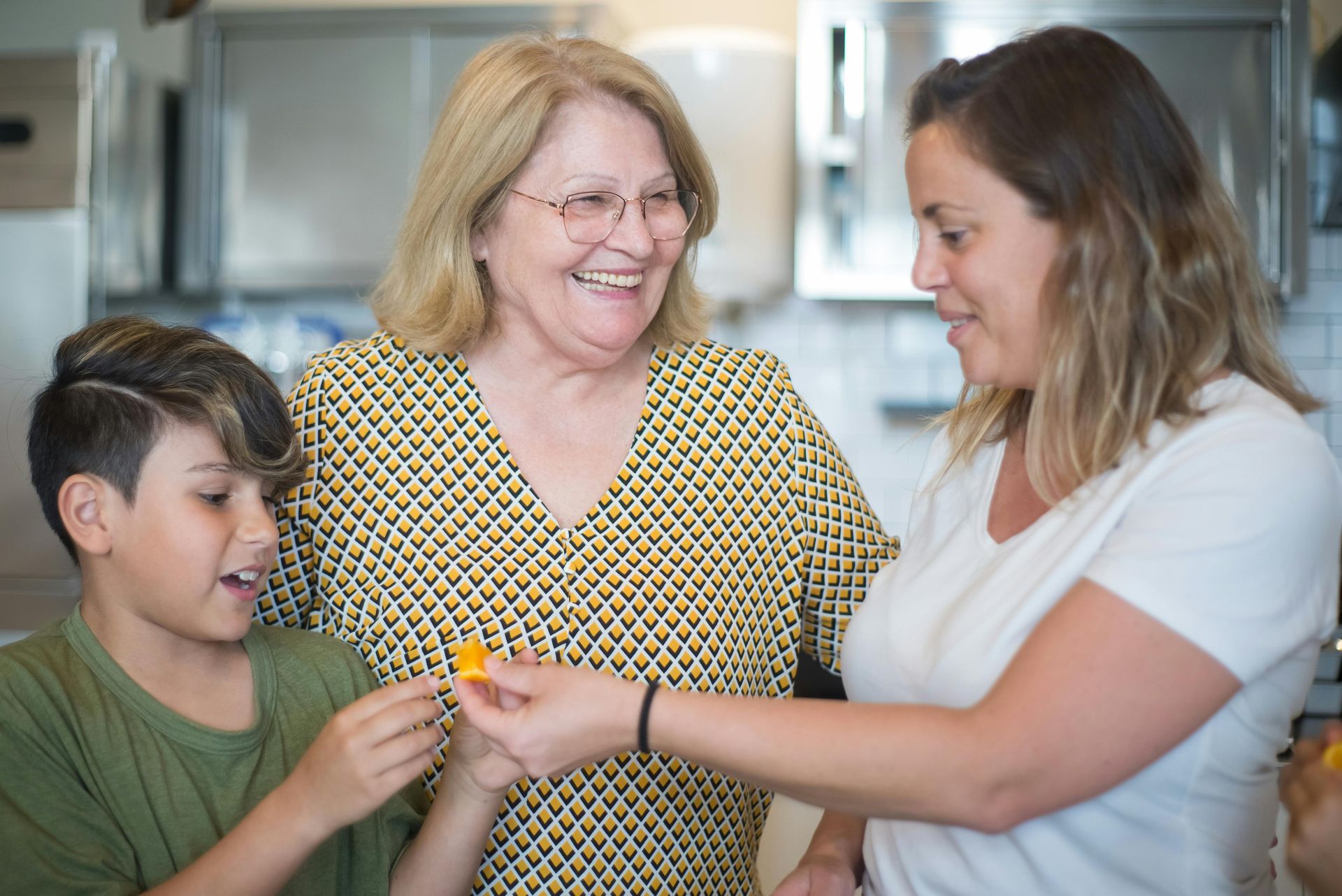 An older woman and a younger person look at a small orange piece of food held by a woman in a kitchen setting.