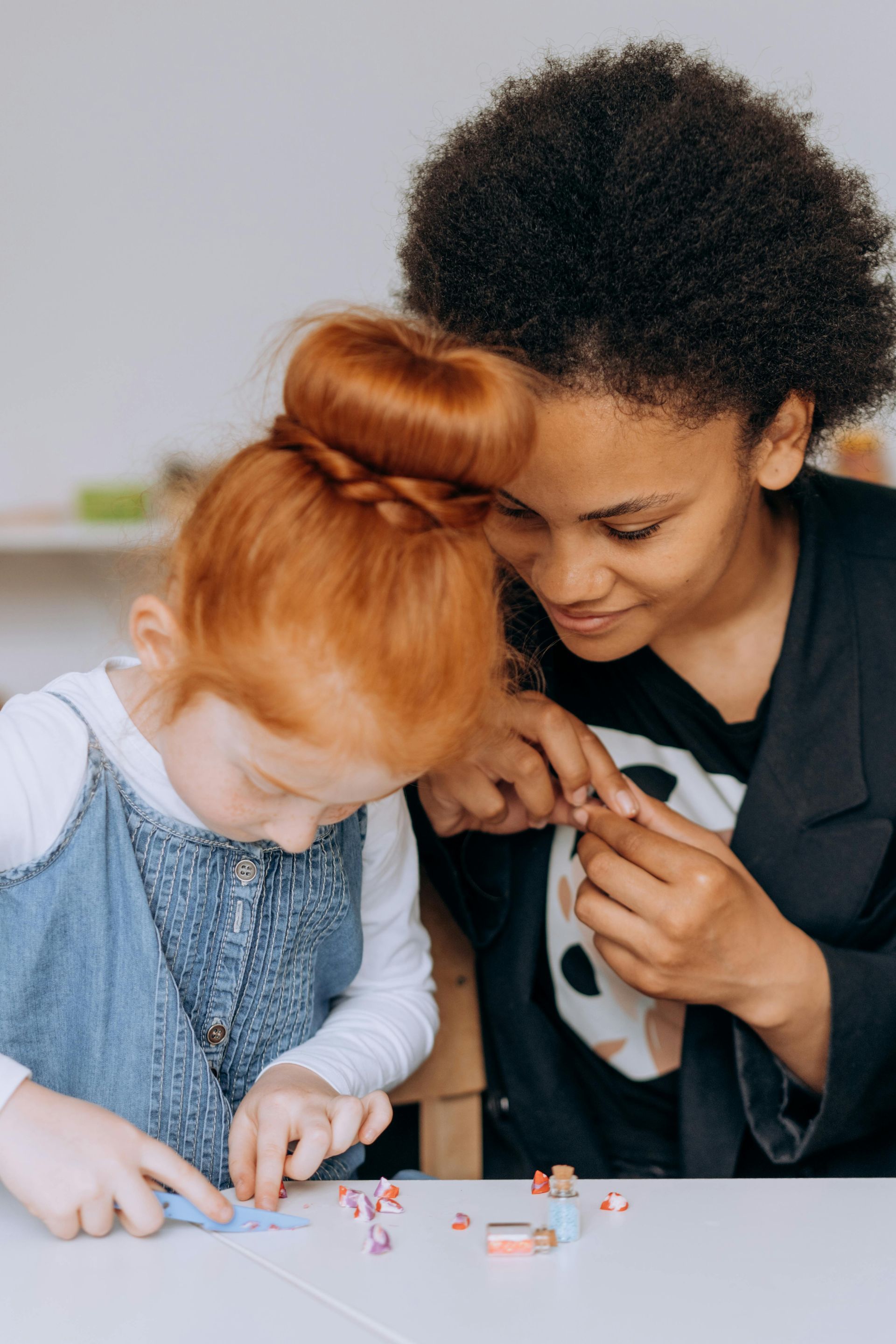 A person with short, dark, curly hair helps a child with a red hair bun work with small craft pieces at a table.