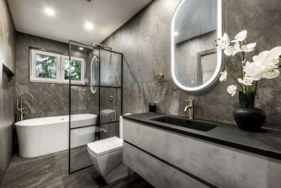 Modern bathroom with gray tile walls and floors. A white tub, toilet, and vanity sit in the space, with gold fixtures and a backlit mirror.
