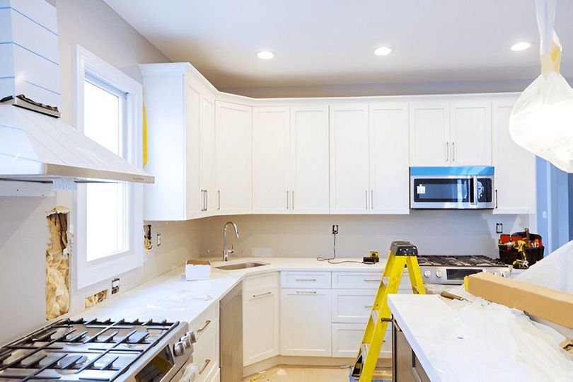 Kitchen under renovation, featuring white cabinets, a stove, and a microwave. A yellow ladder and other construction materials are present.