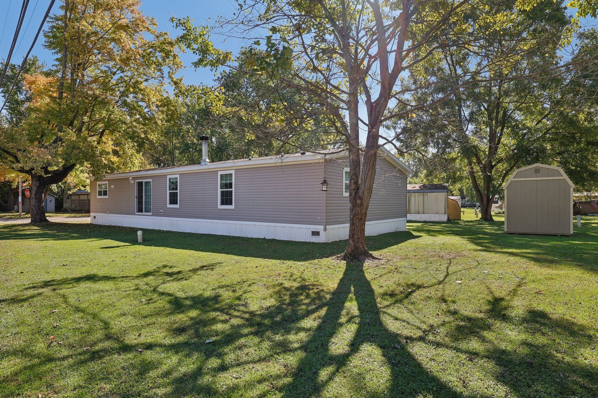 Single-story house with gray siding, shed, and trees on a sunny, grassy lot.
