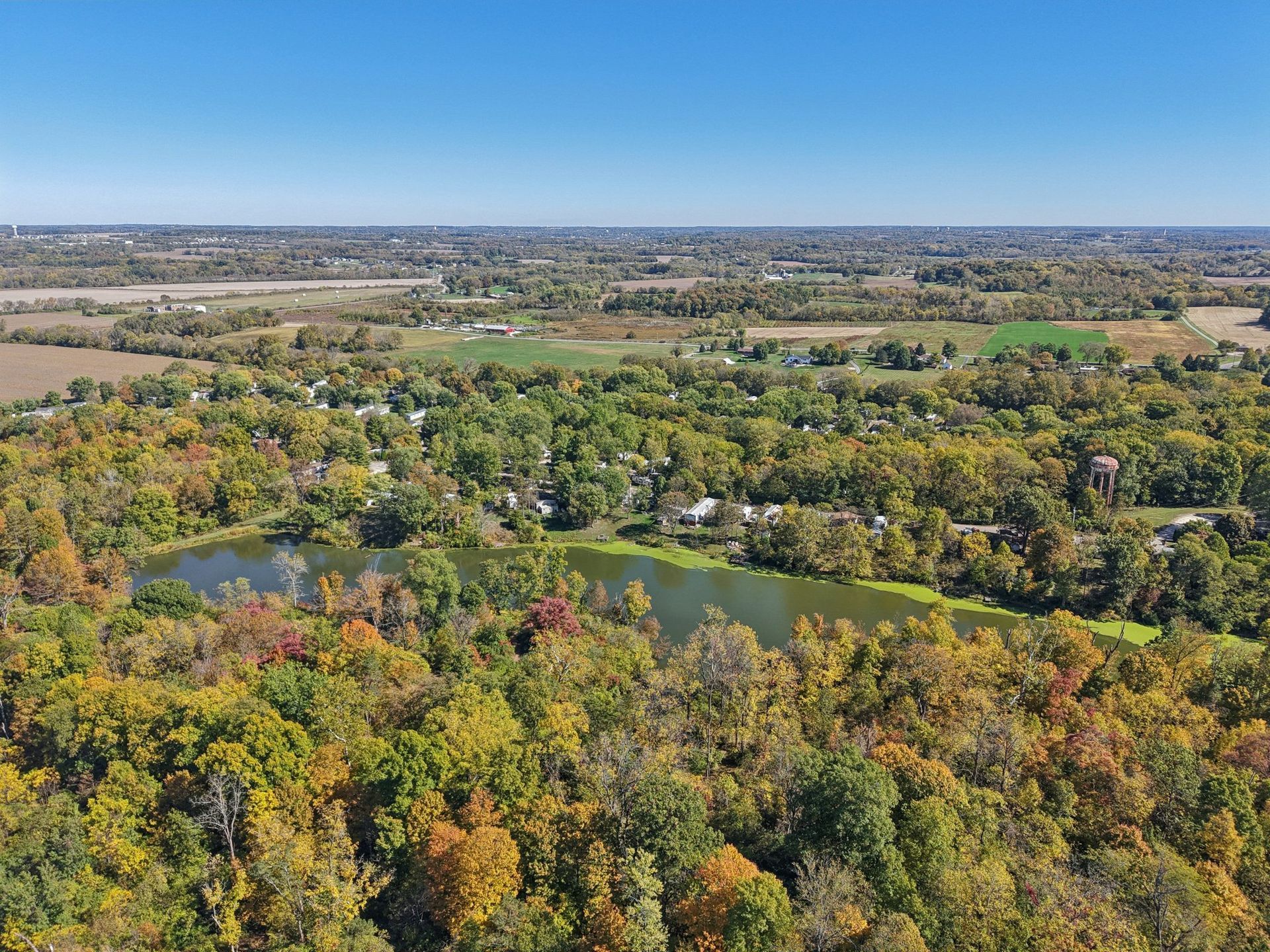Aerial view of a lake surrounded by colorful autumn trees and fields under a blue sky.