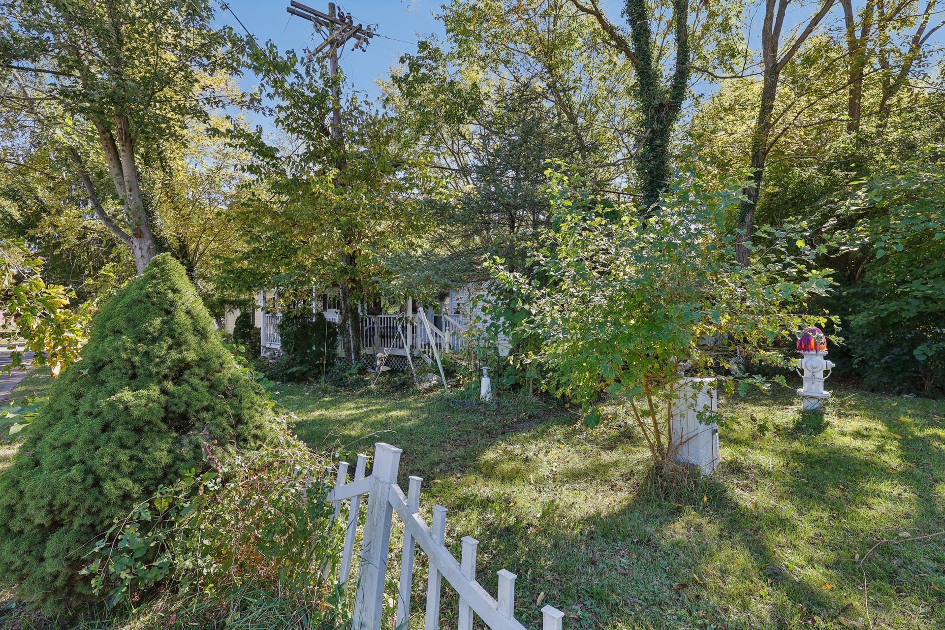 White picket fence leads into a yard with greenery, trees, and a small decorative statue.