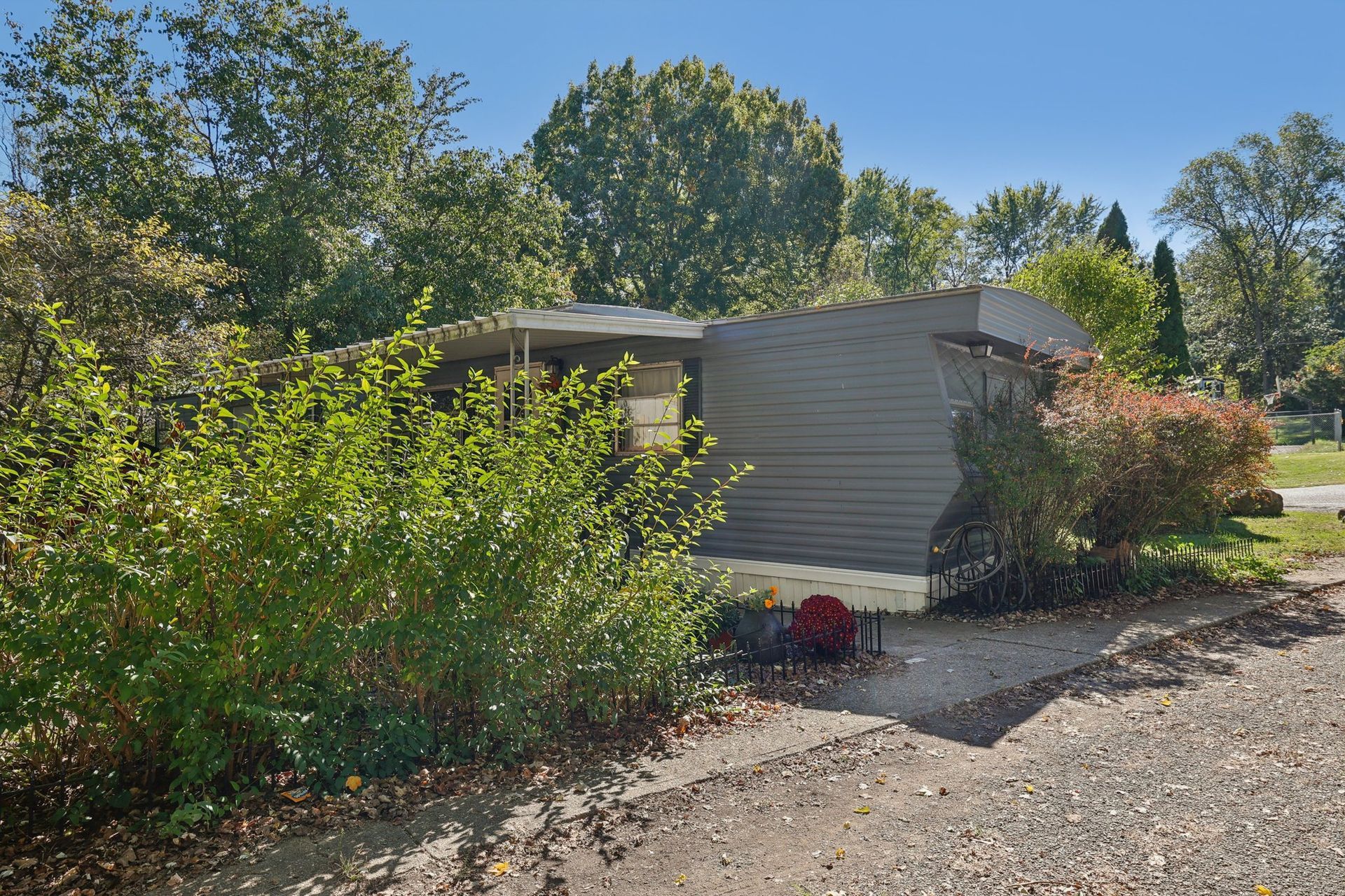 Mobile home surrounded by trees and bushes; gravel driveway in front.