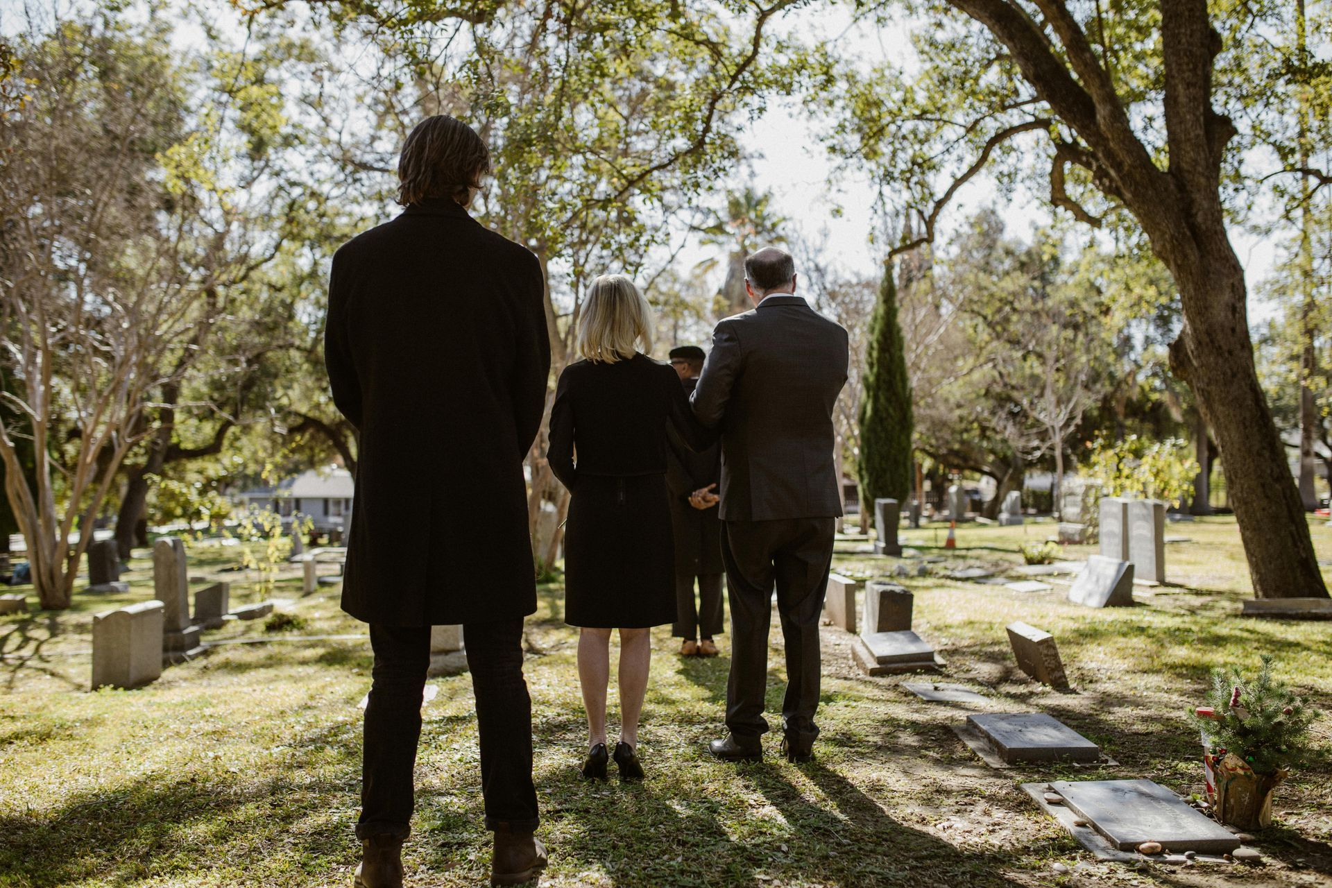 People walking in a cemetery, possibly a funeral. Group is in dark attire, trees and tombstones surround them.