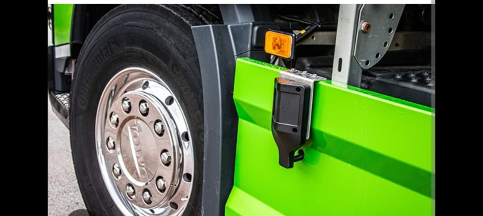 Primo piano di una ruota e di un parafango verdi di un camion con un sensore nero attaccato. È visibile un cartello di pericolo biologico.