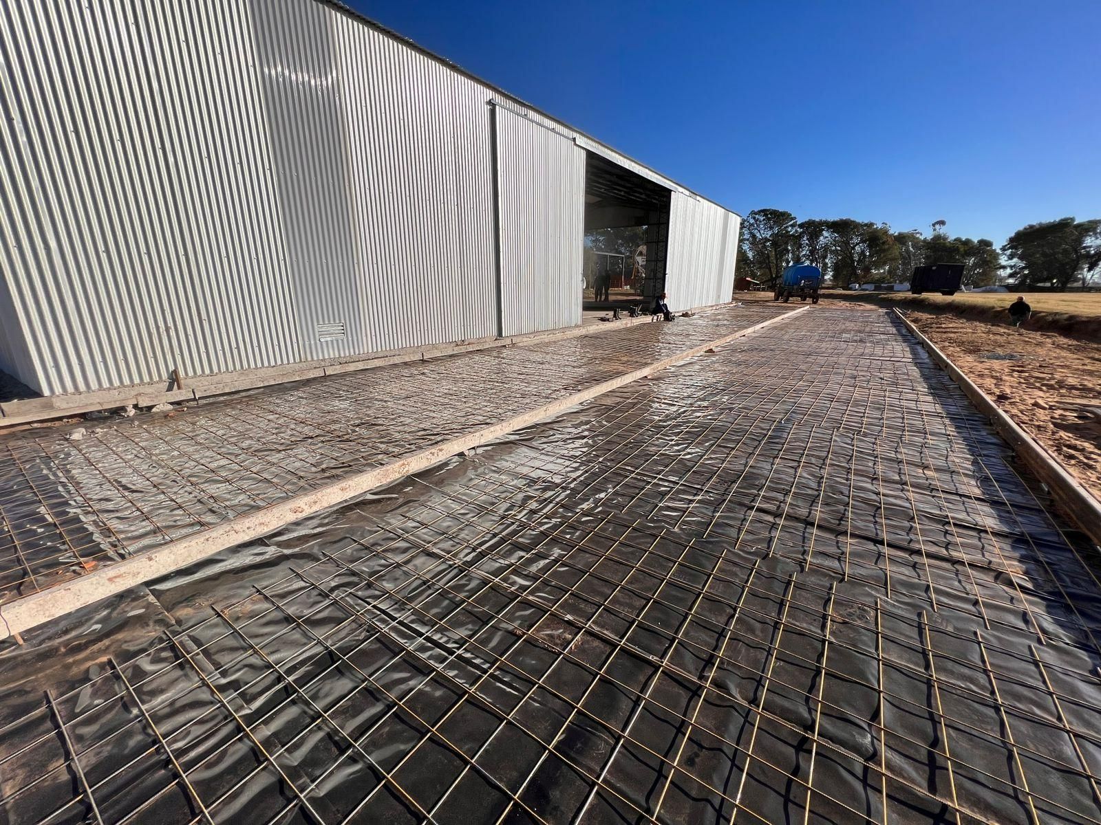 Se vierte una nueva losa de hormigón frente a un edificio de chapa ondulada. Se ve la malla de refuerzo.