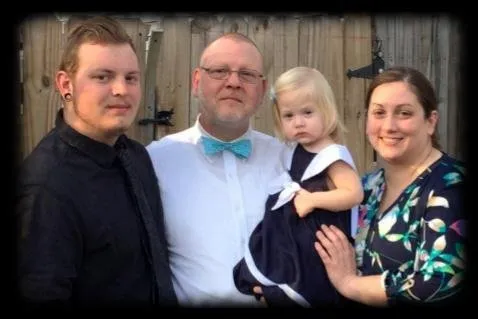 Family of four posing outdoors, smiling. Man in blue bow tie holds young girl in dress. Two adults, one young adult.