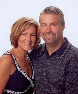 Smiling couple posing for a photo. Woman in blue top, man in a blue shirt, both in front of a white backdrop.