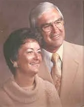 Couple smiling; man in suit, glasses, white hair; woman in sweater, dark hair, earrings.