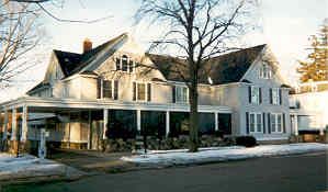 Two-story pale house with dark shutters, covered porch, and snow on the ground.