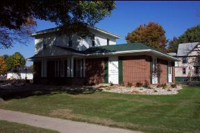 A two-story building with a brick addition and green roof on a grassy lawn with trees.