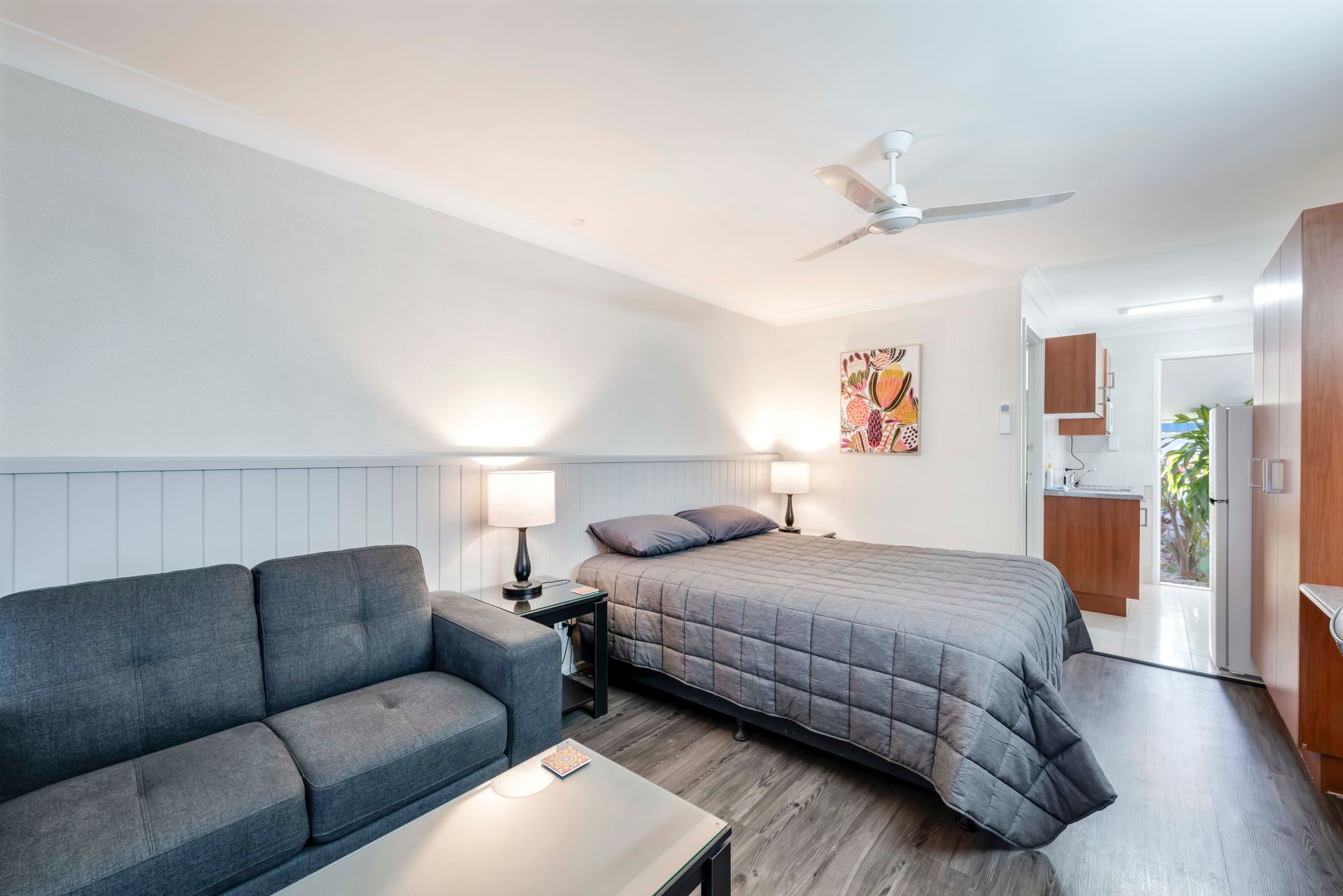 Bright bedroom with twin beds, desk, chairs, and blue accents in a compact hotel-style room.