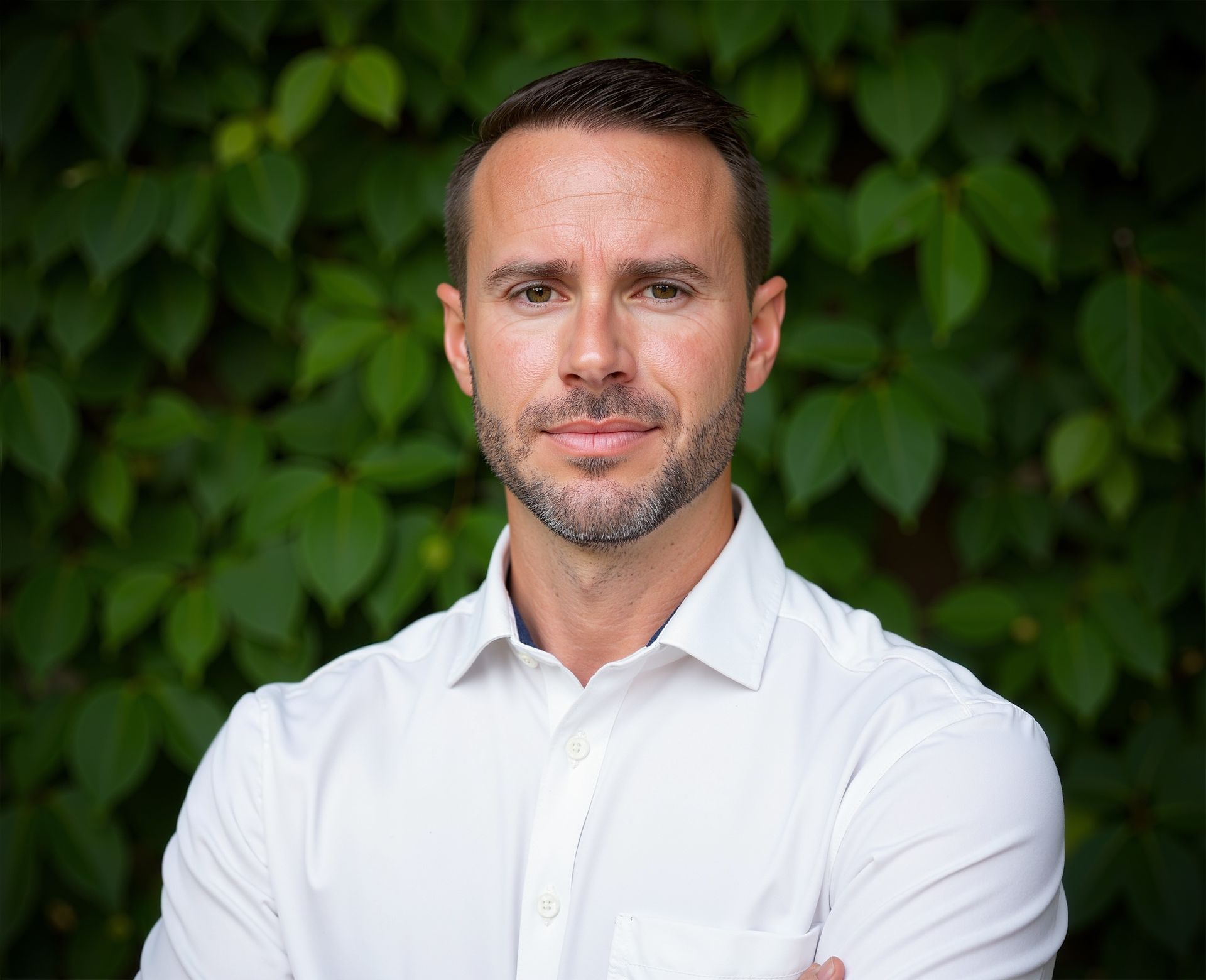 Man in white shirt, arms crossed, smiling. Green leafy backdrop.