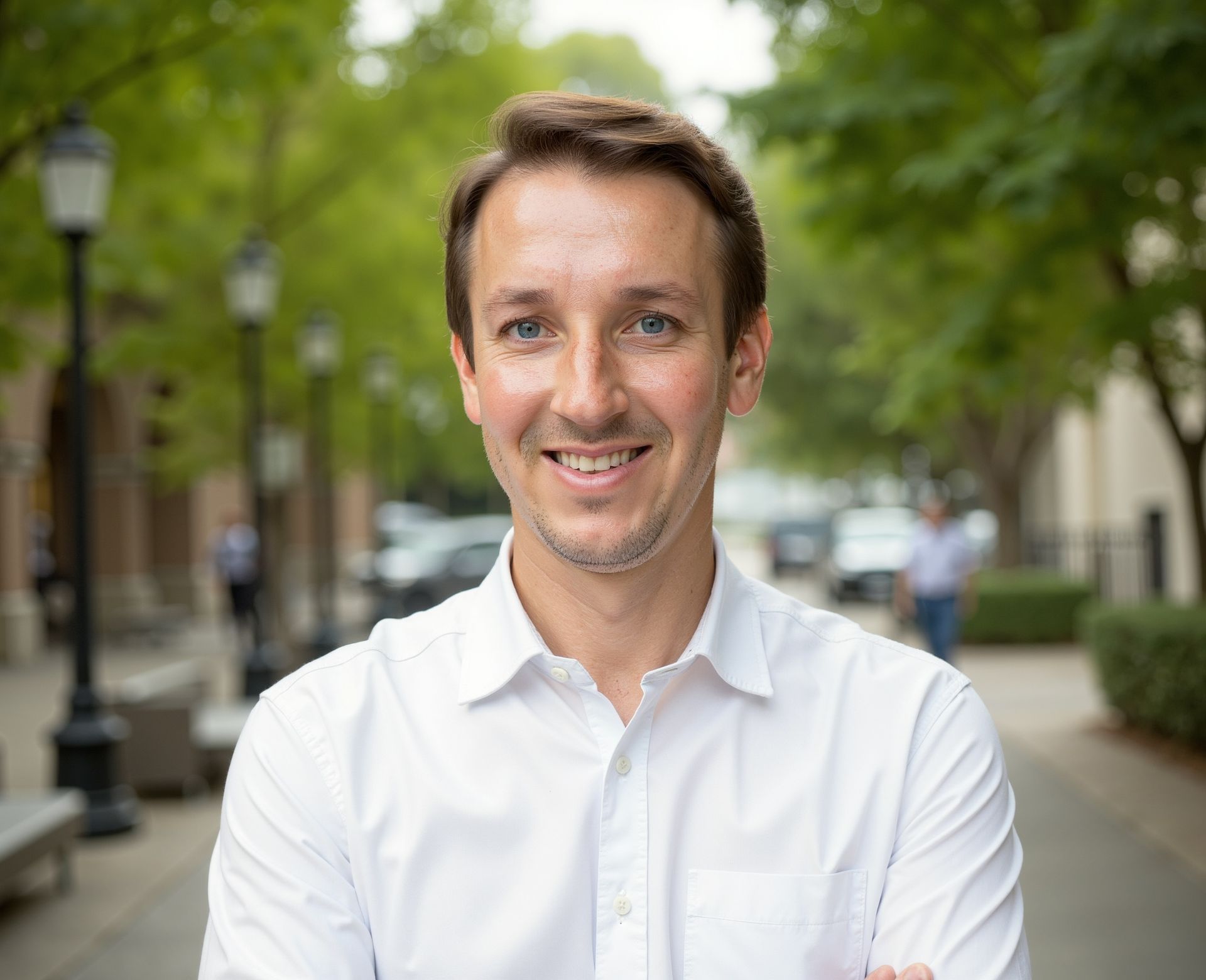 Man in white shirt smiles outdoors, blurred background of street and trees.