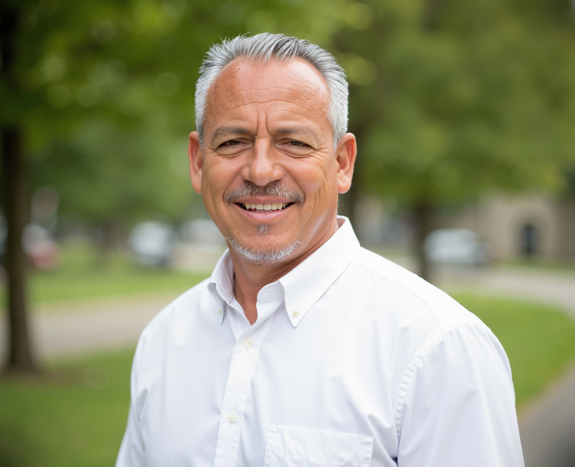 Man with gray hair and a white shirt smiling outdoors, trees in background.