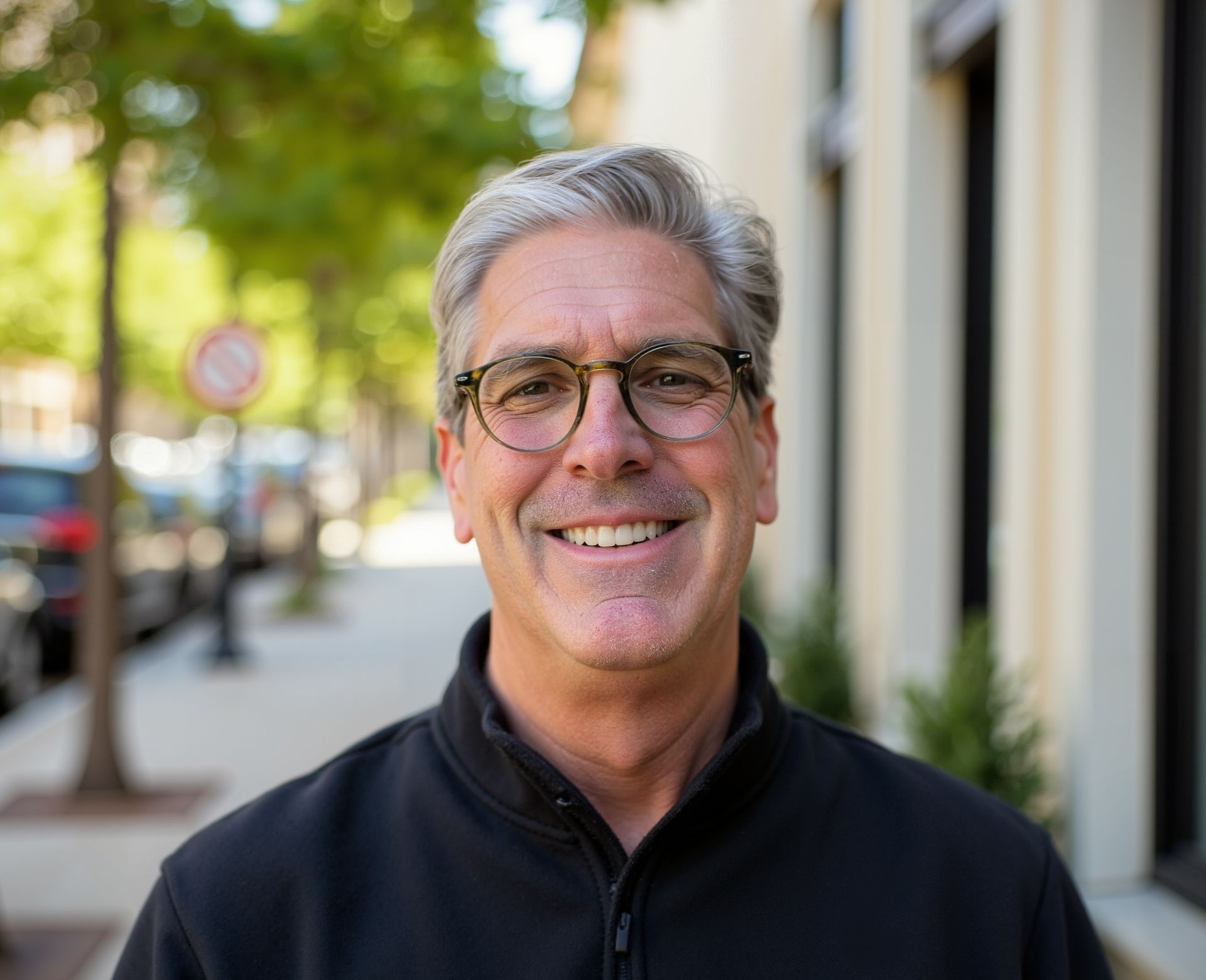 Man with glasses smiling outdoors, wearing black jacket, blurred background of street and building.