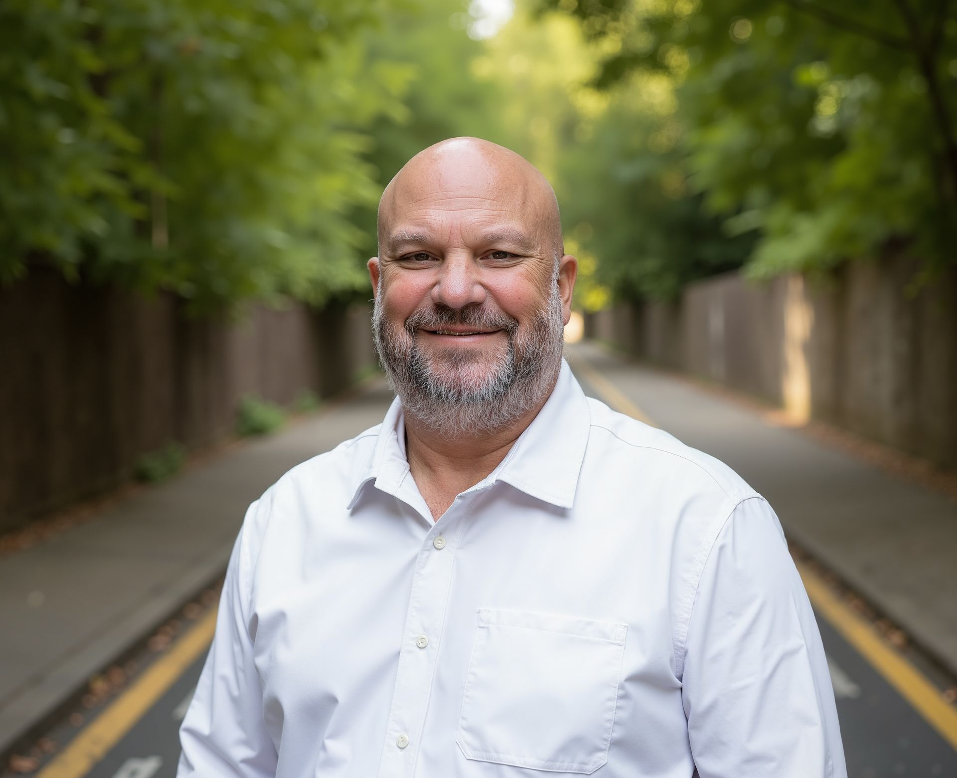 Man with beard, bald head, in white shirt, smiling outdoors on a road lined with trees.