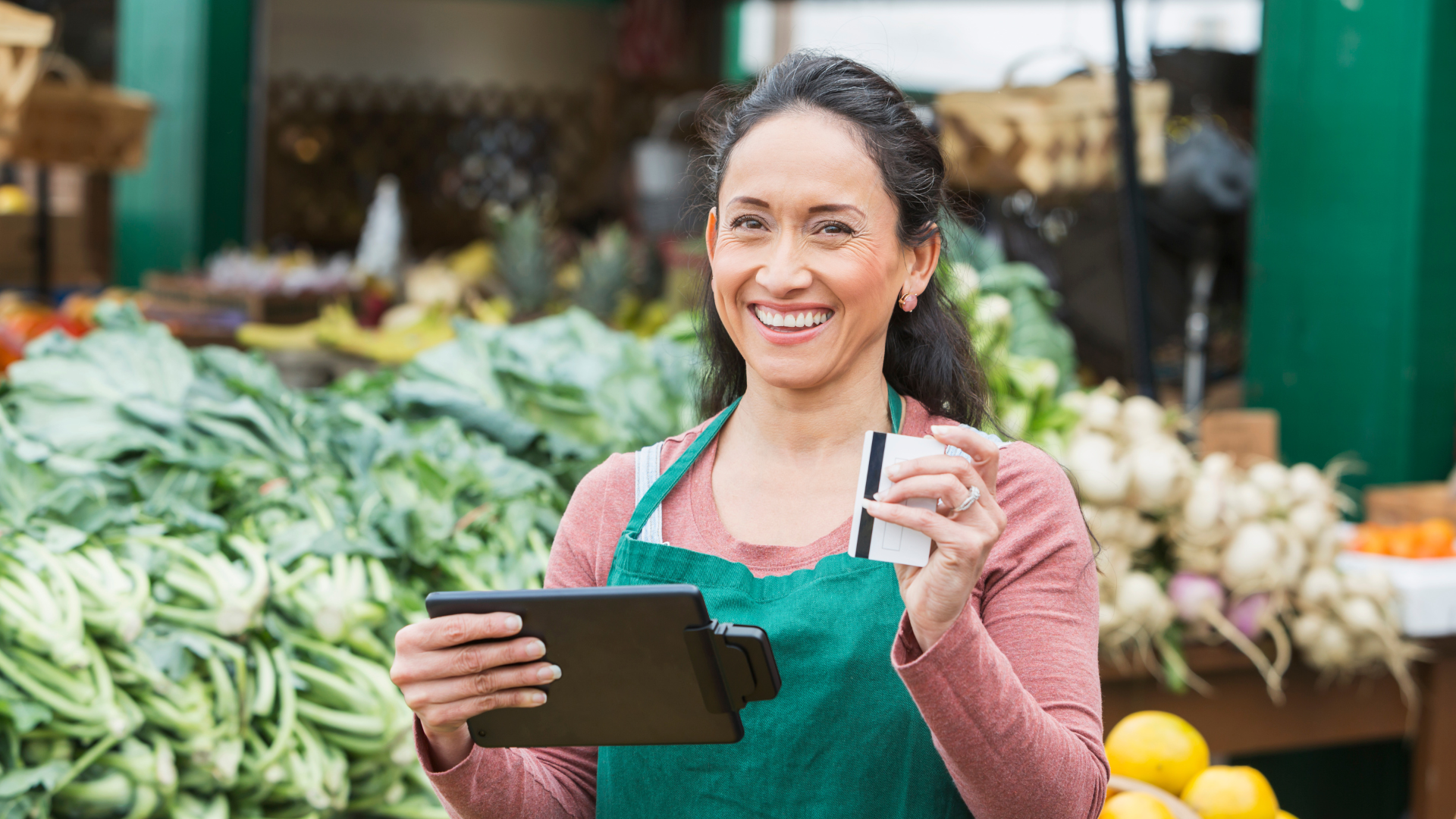 A woman is standing in front of a vegetable stand holding a tablet and a credit card.