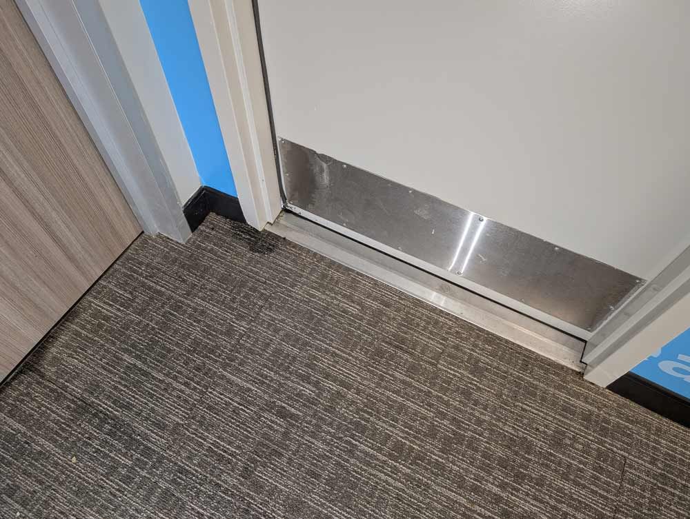 A door with a metal kick plate opens into a room with gray carpet and blue walls.