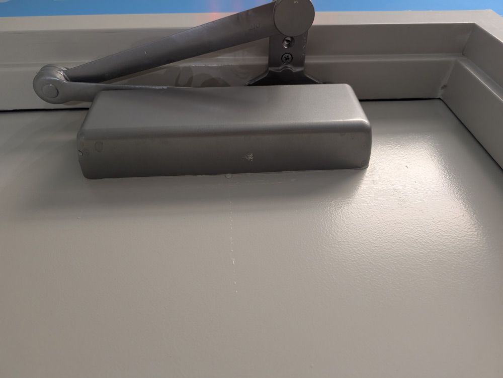 A gray hydraulic door closer mounted on the upper portion of a white door shows water leaking inside.