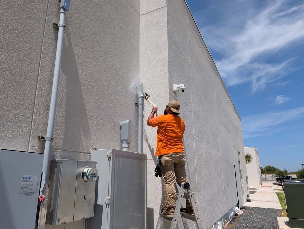 A person in an orange shirt standing on a ladder, using a hose and water on the exterior wall of a building.