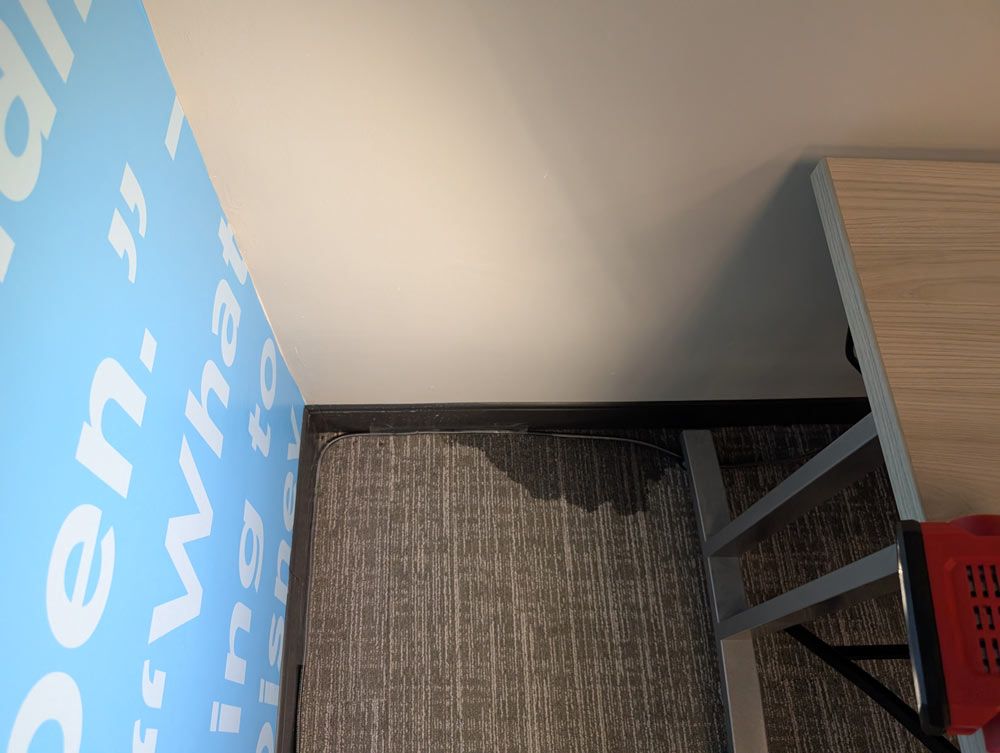 A damp, dark stain on gray patterned office carpet near a wall with blue branding and a desk leg.