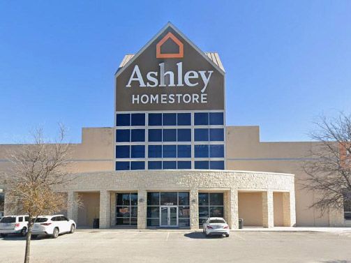 The storefront of an Ashley Homestore with a large tan building and glass entrance, viewed from a parking lot.