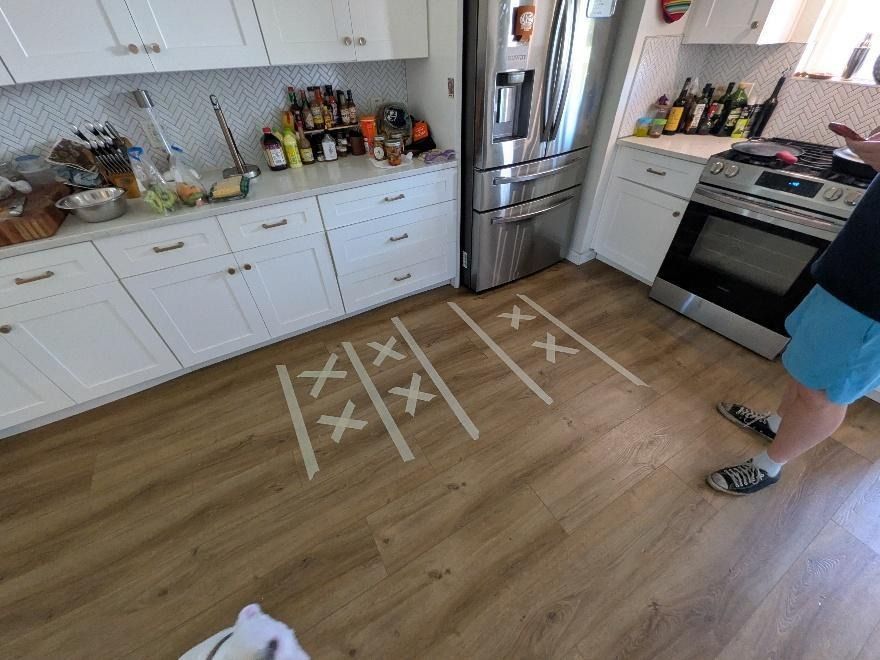 Kitchen floor with taped X marks; a person stands near the stove and refrigerator.