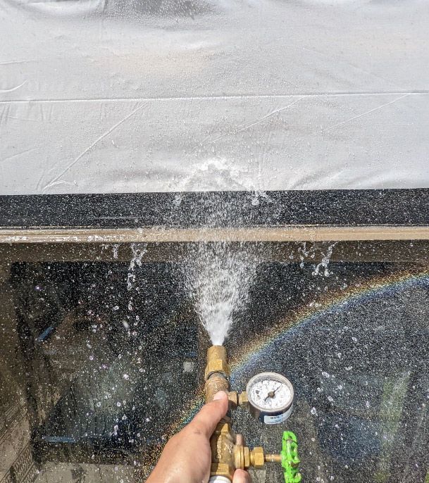 Hand holding hose nozzle spraying water at a wall, with gauge and rainbow visible