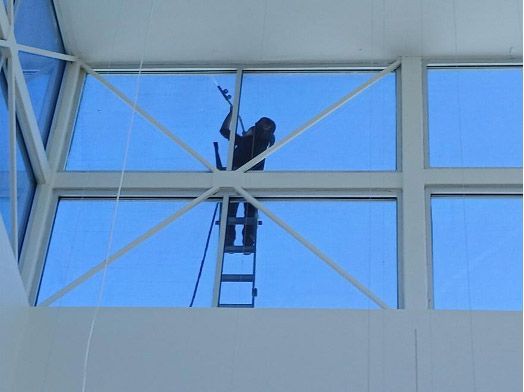 A worker on a tall ladder uses a squeegee to clean a high, windowed wall with white framing against a bright blue sky.