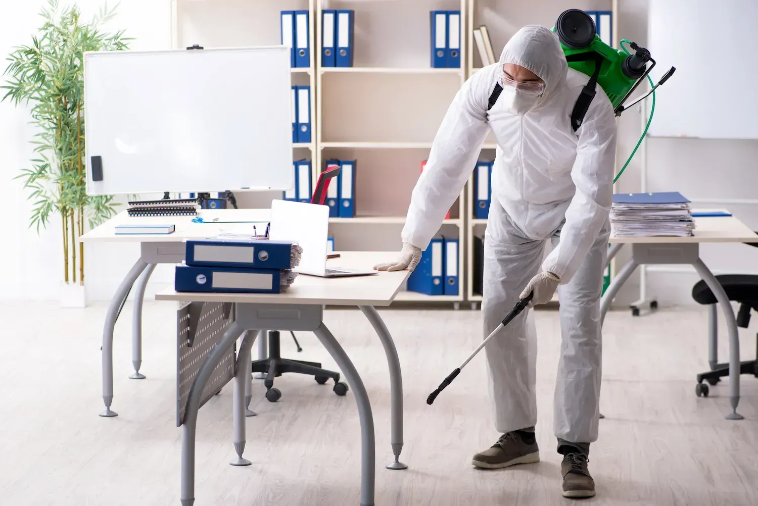 A man in a protective suit is disinfecting an office with a sprayer.