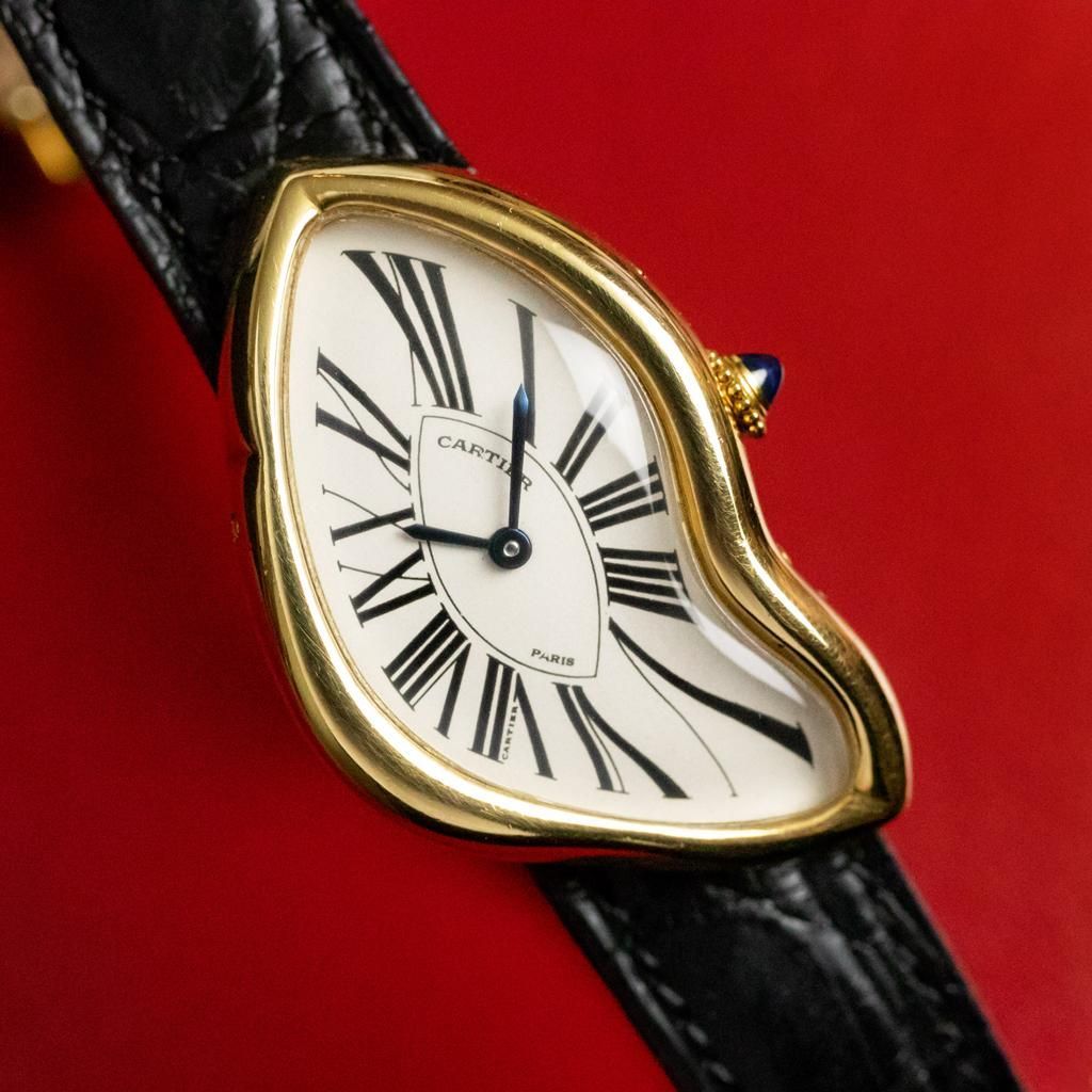 Gold Cartier wristwatch with an asymmetrical, melting-style case, black strap, and Roman numeral dial.