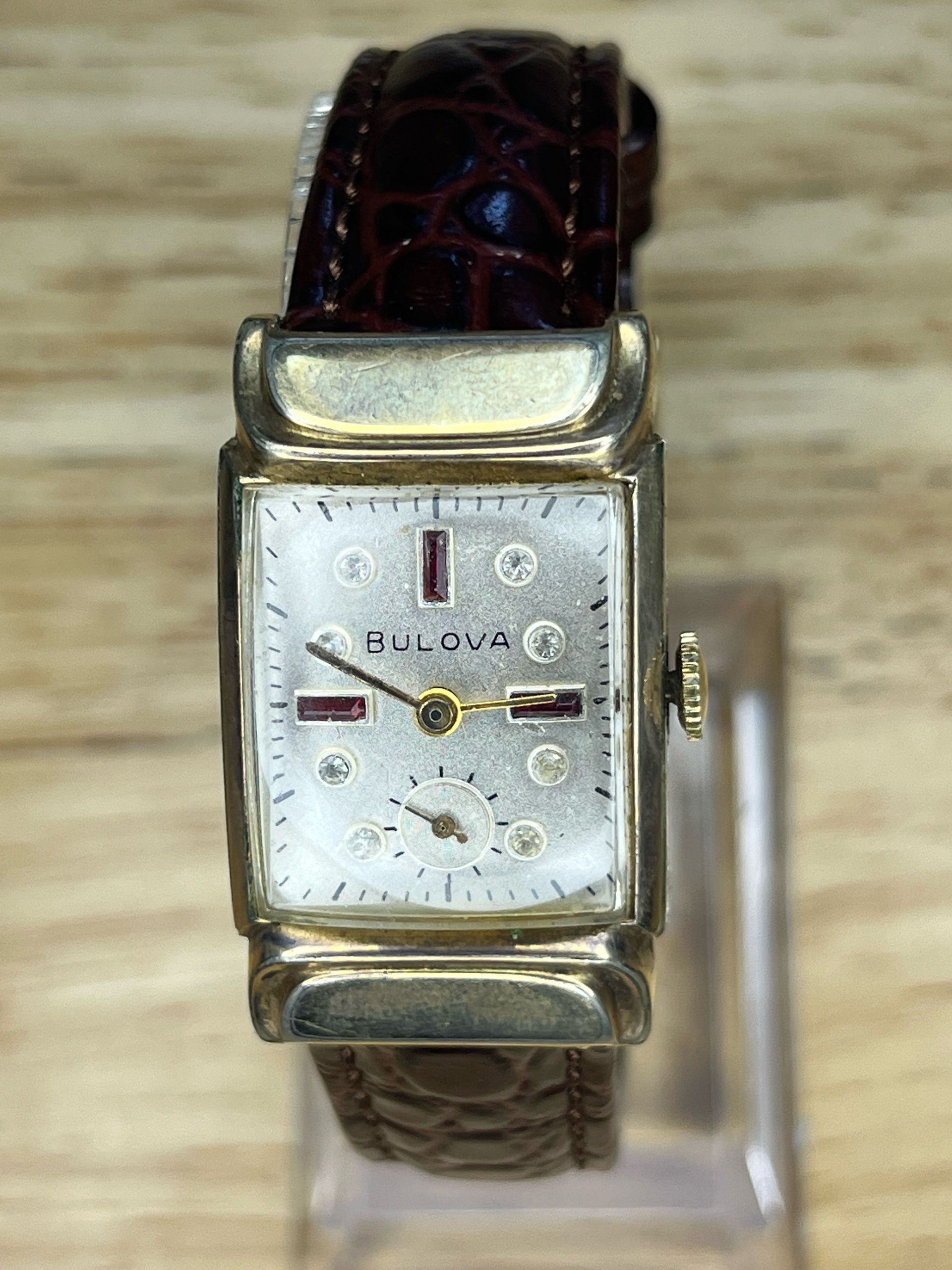 Gold-tone wristwatch with a rectangular face, small second dial, and brown leather strap on a stand.