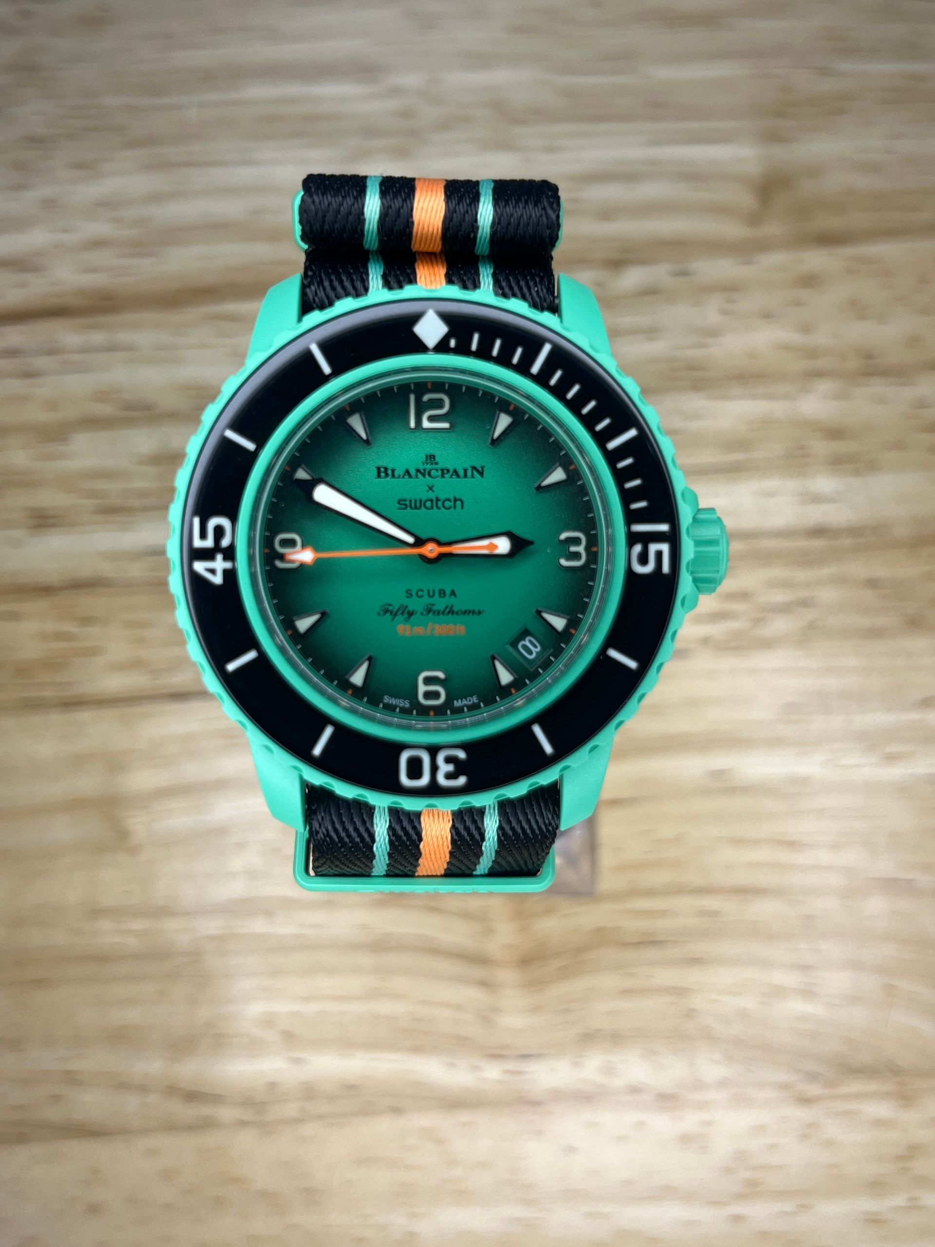 Green and black wristwatch with a teal bezel and a black and orange striped band on a wood surface.