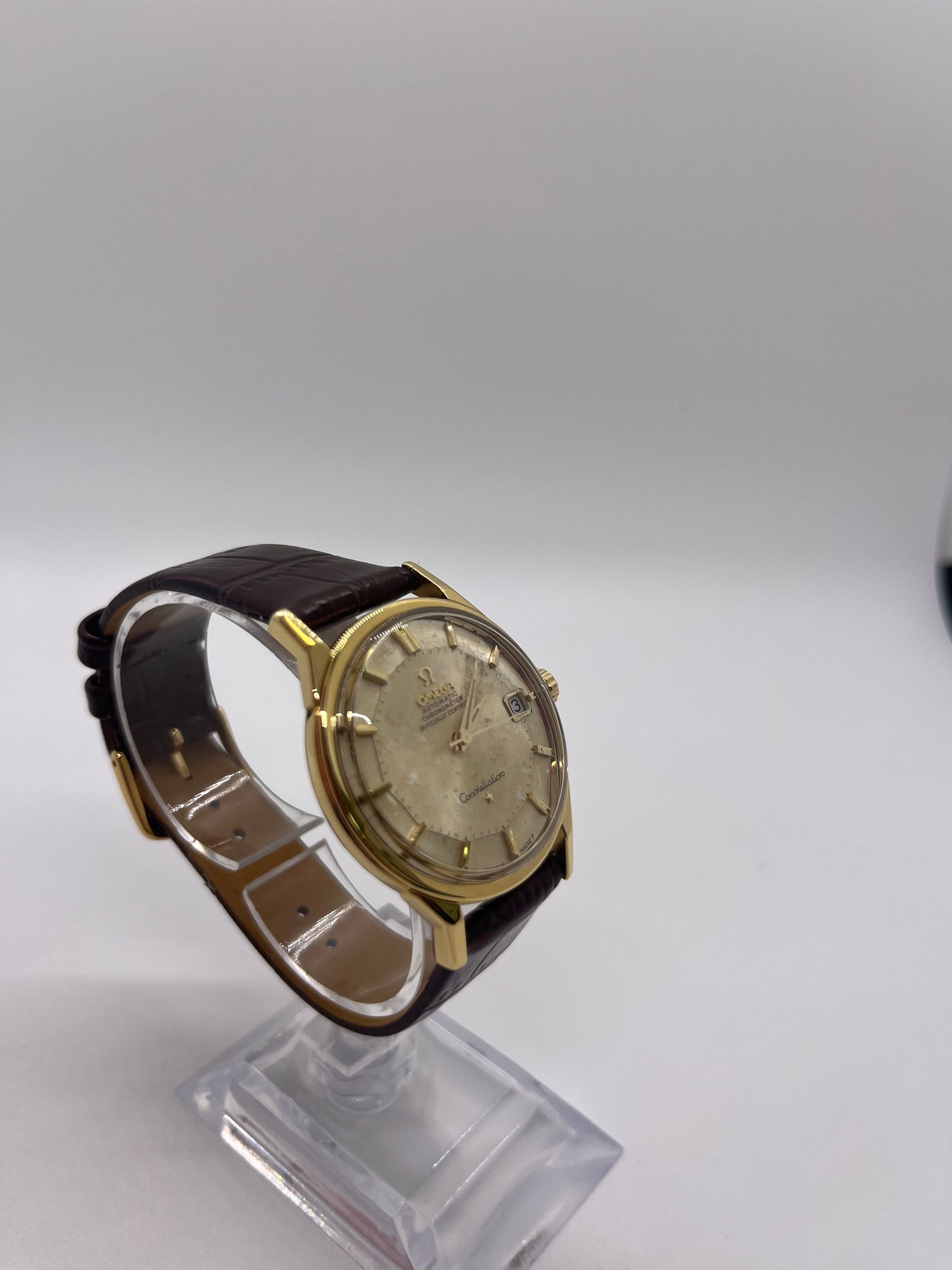 Gold-toned vintage wristwatch on a clear stand; brown leather band, beige face with date window.