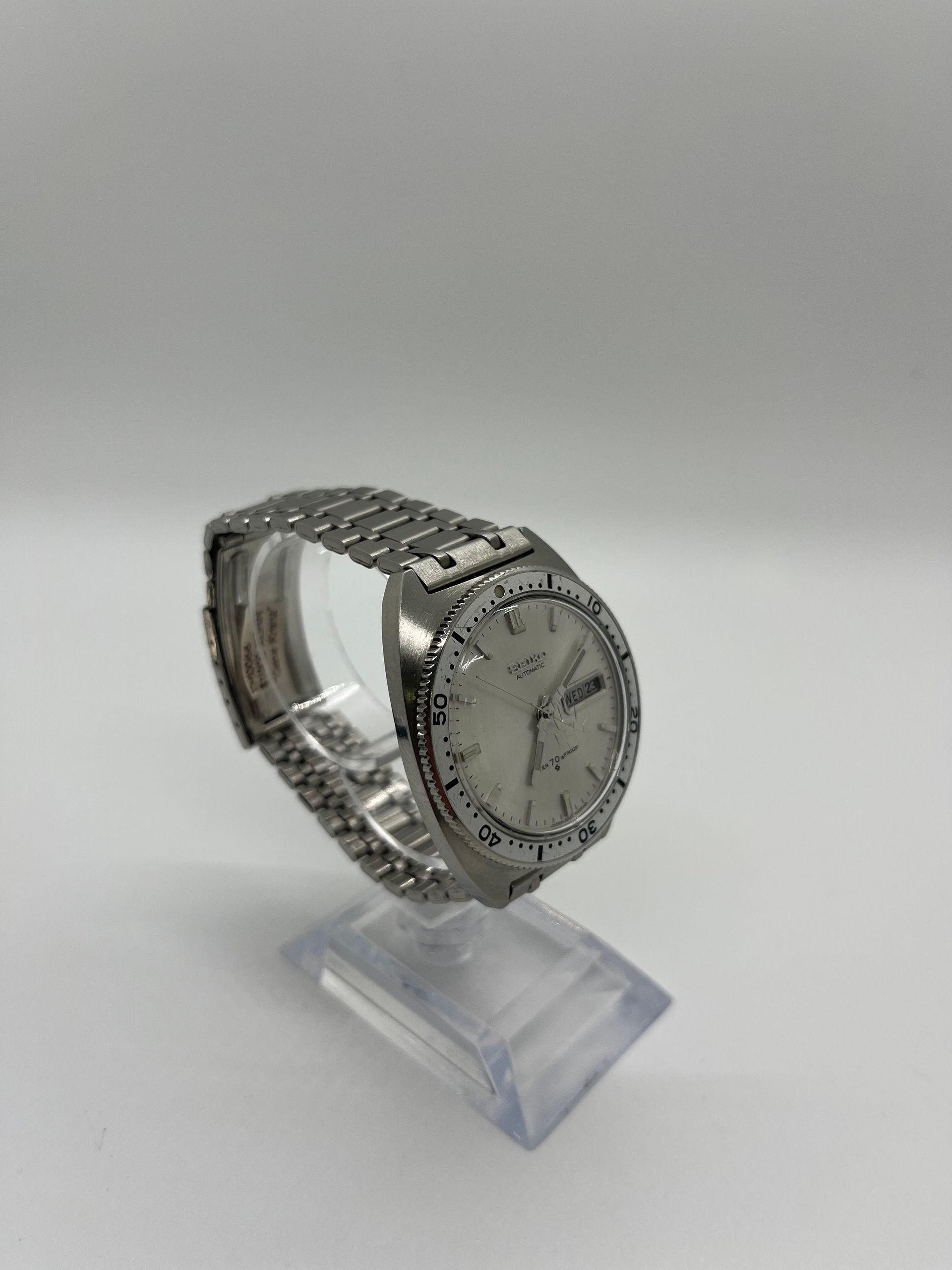 Silver wristwatch with a day/date display on a clear stand, against a white background.