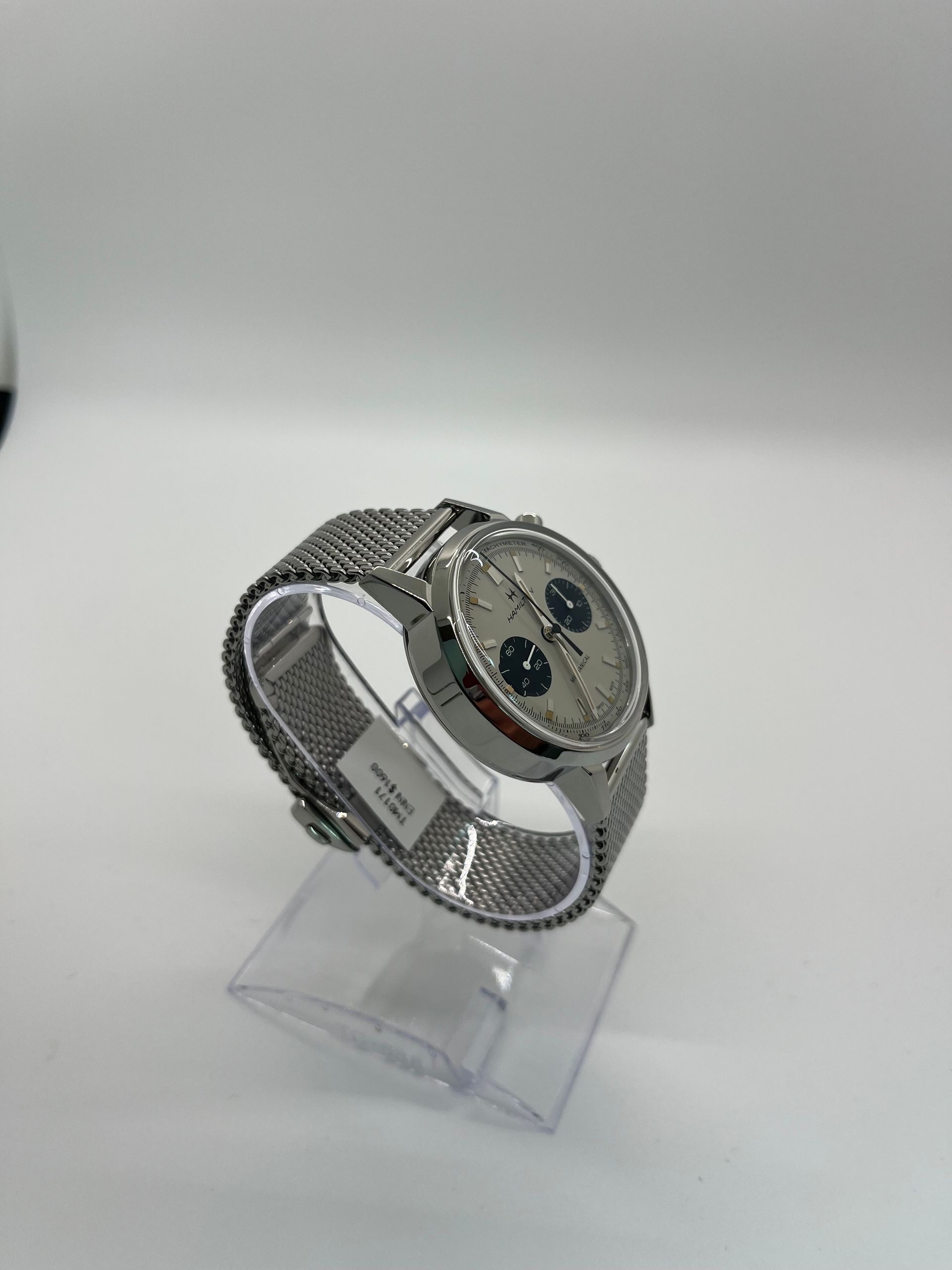 Silver Breitling watch with a mesh band, on a clear stand, against a white backdrop.