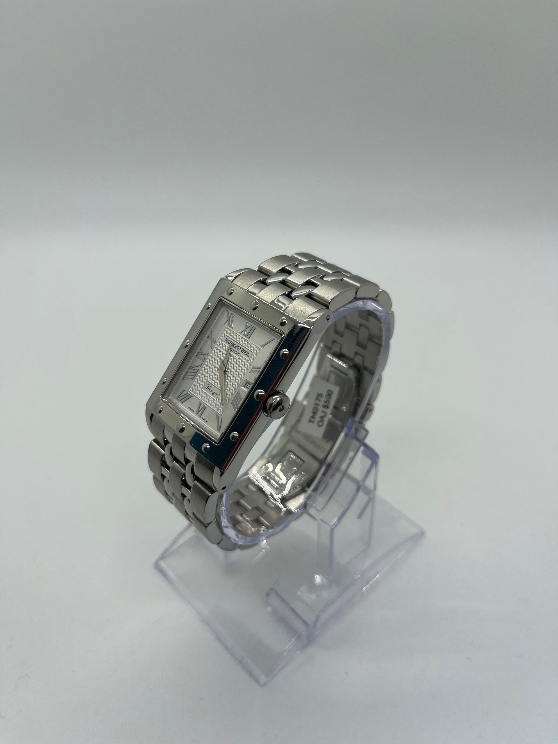 Silver rectangular Tiffany watch on a clear stand. White face with Roman numerals, silver band.