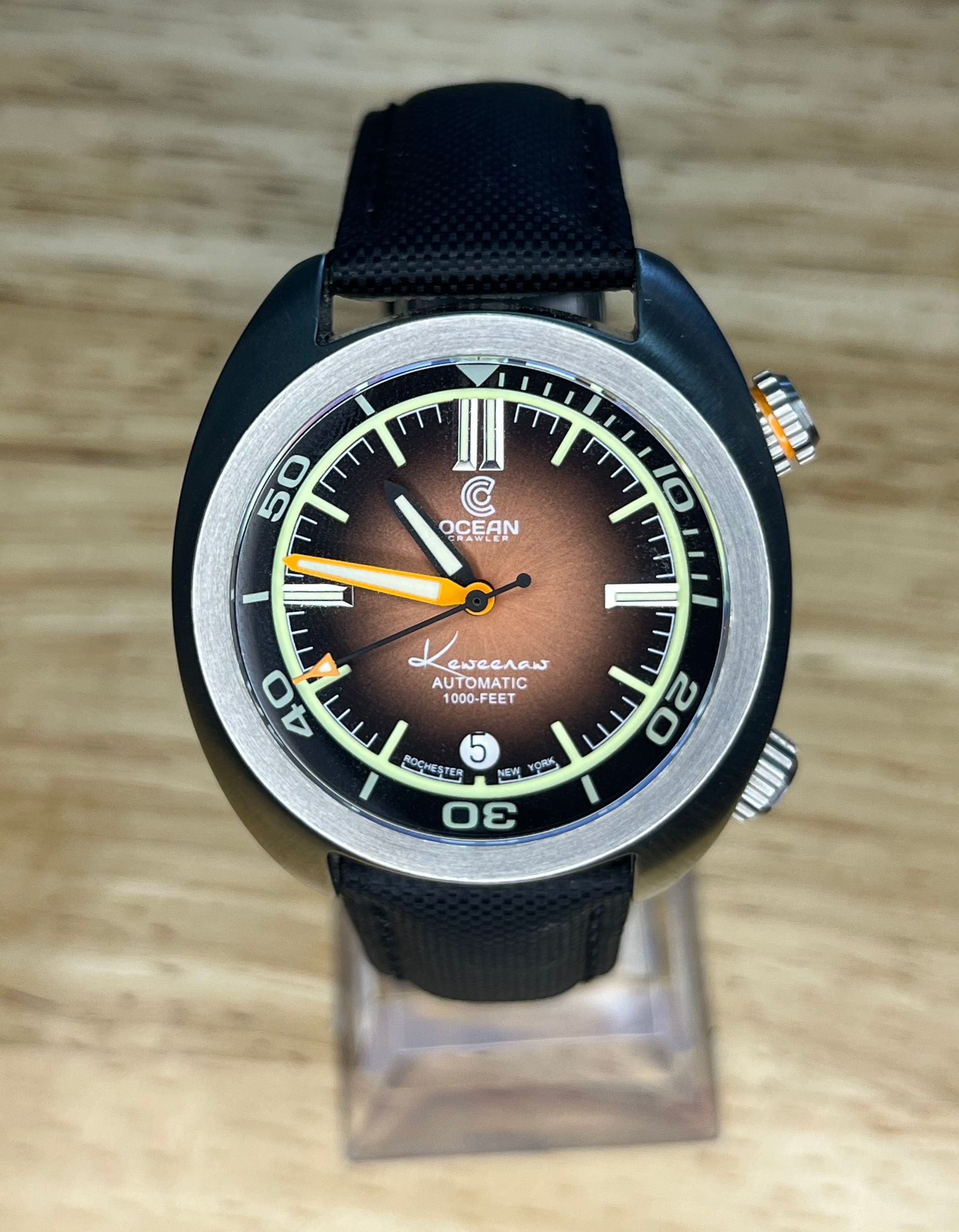 Black and silver wristwatch with a brown face and orange accents on a clear stand.