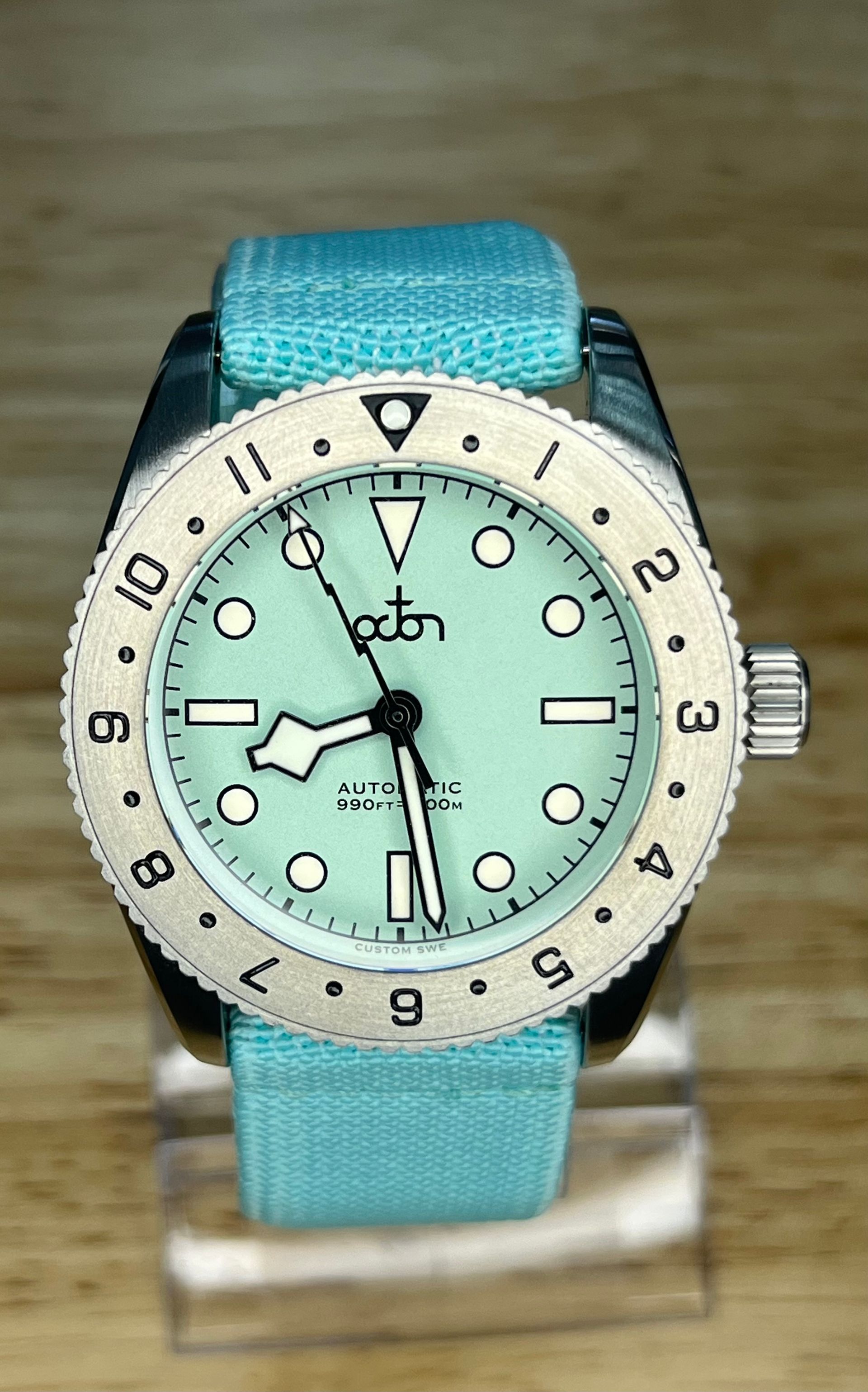 A wristwatch with a teal face and bezel, on a matching teal fabric strap, displayed on a wood surface.