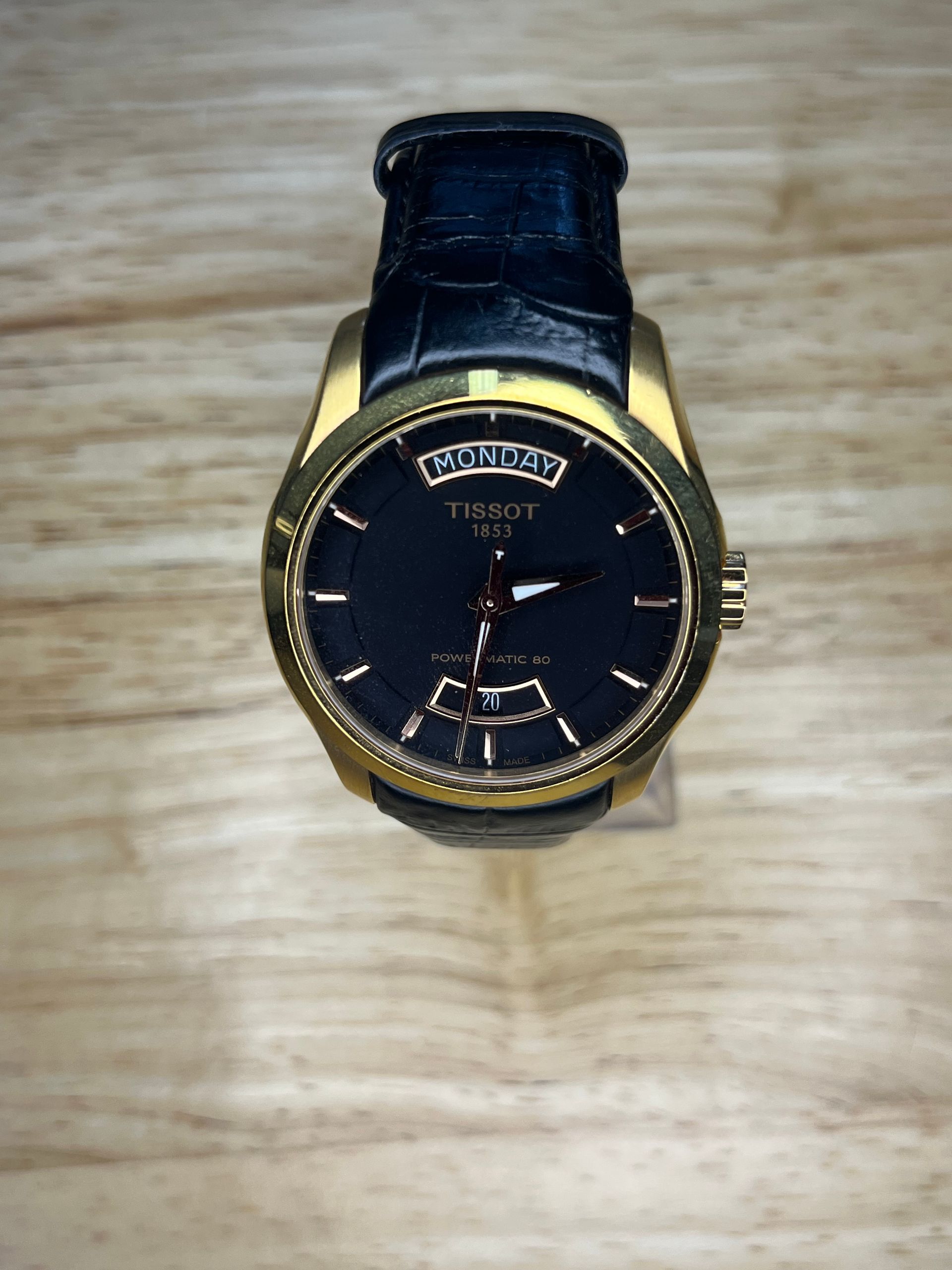 Gold Tissot watch with a black face and a black leather strap on a wooden surface.