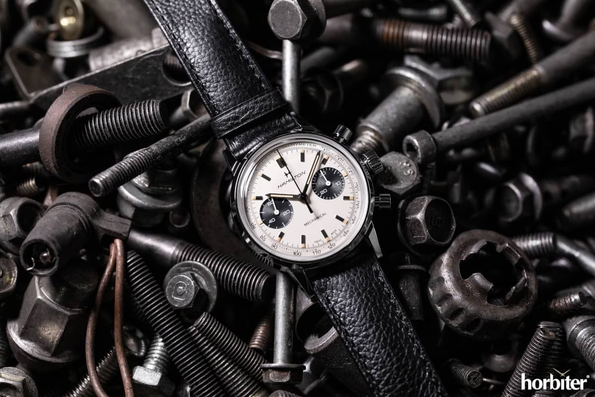 A vintage wristwatch with a black leather band rests on a pile of metal bolts and hardware.